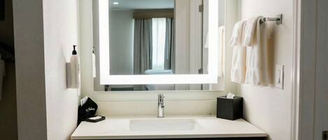 A hotel bathroom with a large mirror, a white sink, a black soap dispenser, and a towel rack with white towels.