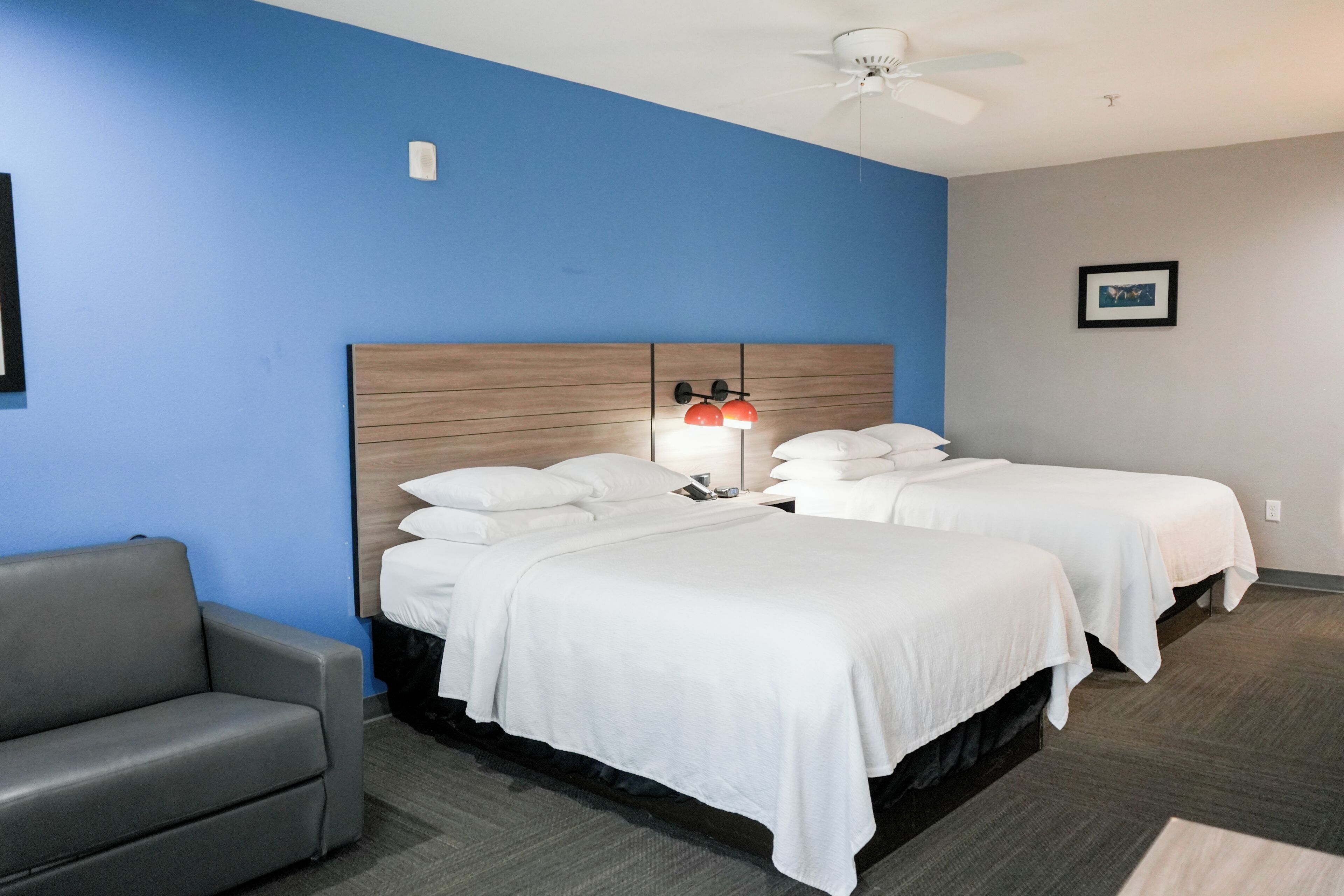 A hotel room with two beds, a gray sofa, a ceiling fan, and a framed picture on the wall.