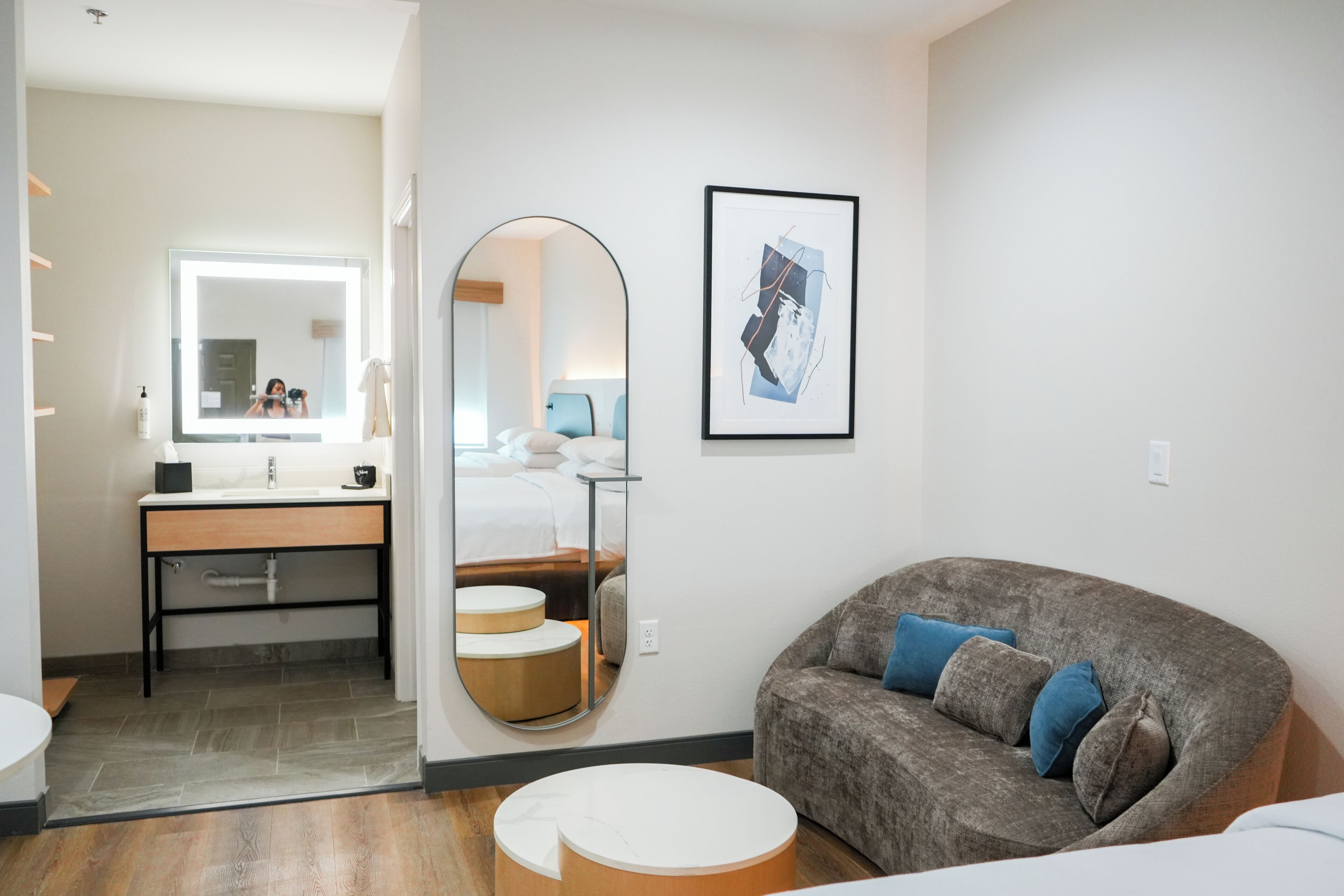 Una habitación de hotel moderna con un espejo grande, una mesa redonda para café, un sofá con cojines y una obra de arte enmarcada en la pared.