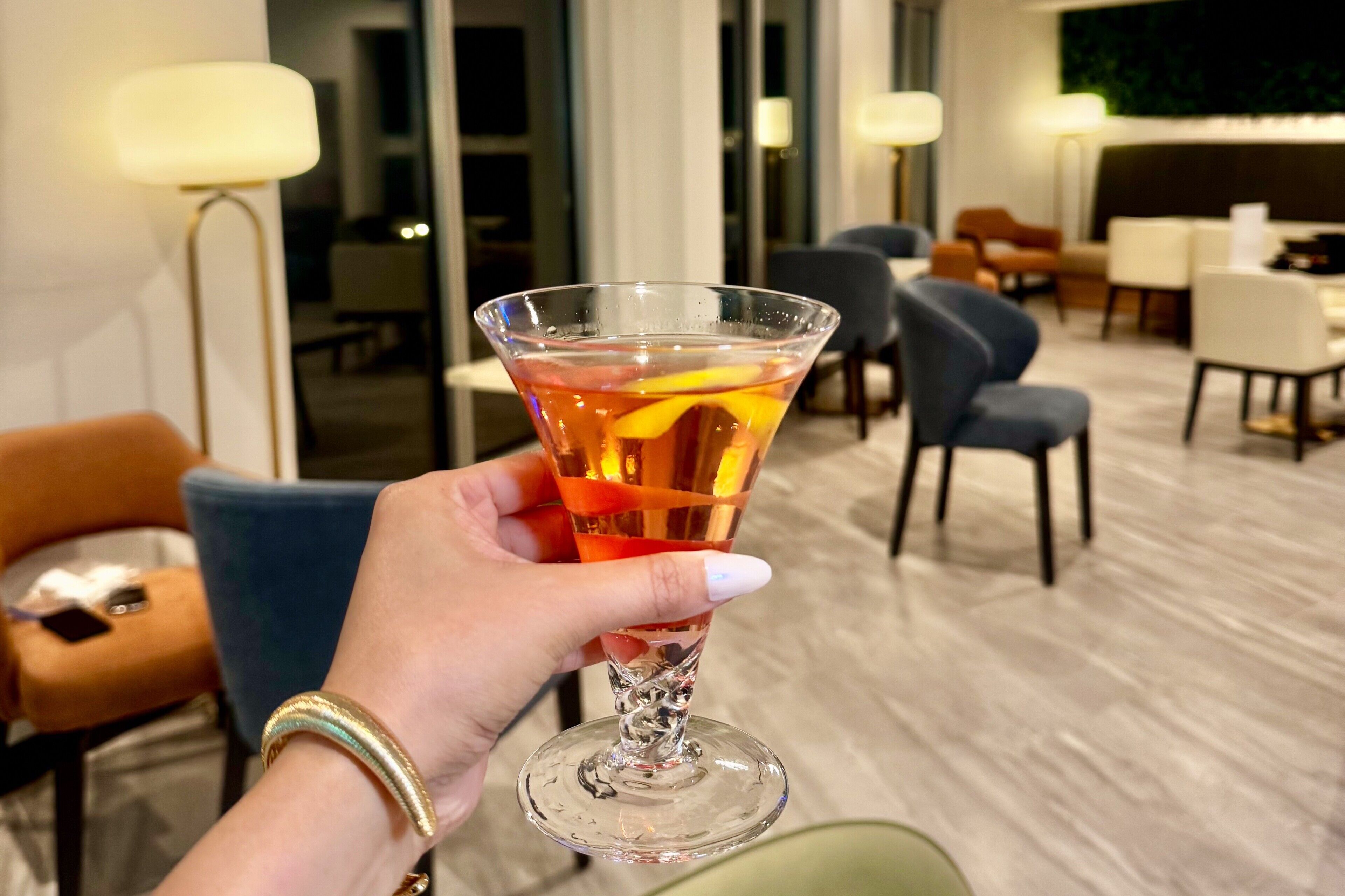 Een hand die vasthoudt een cocktailglas met een drankje en een oranje schijfje, in een moderne hotel lobby met zitplaatsen en lampen.