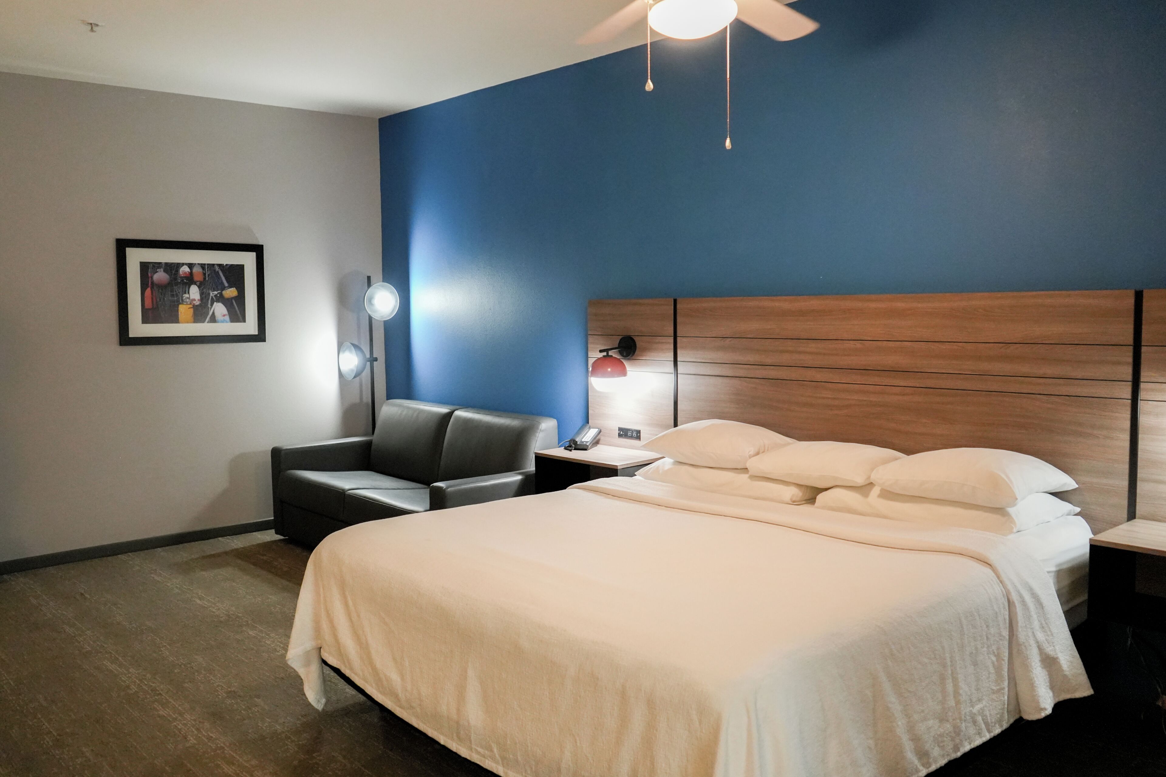 A hotel room with a bed, a desk, a chair, a ceiling fan, and a wall-mounted lamp.