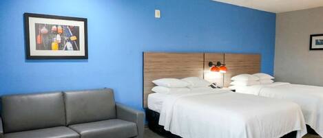 Une chambre d’hôtel avec un mur bleu, un canapé gris, deux lits avec du linge de lit blanc et un ventilateur de plafond.