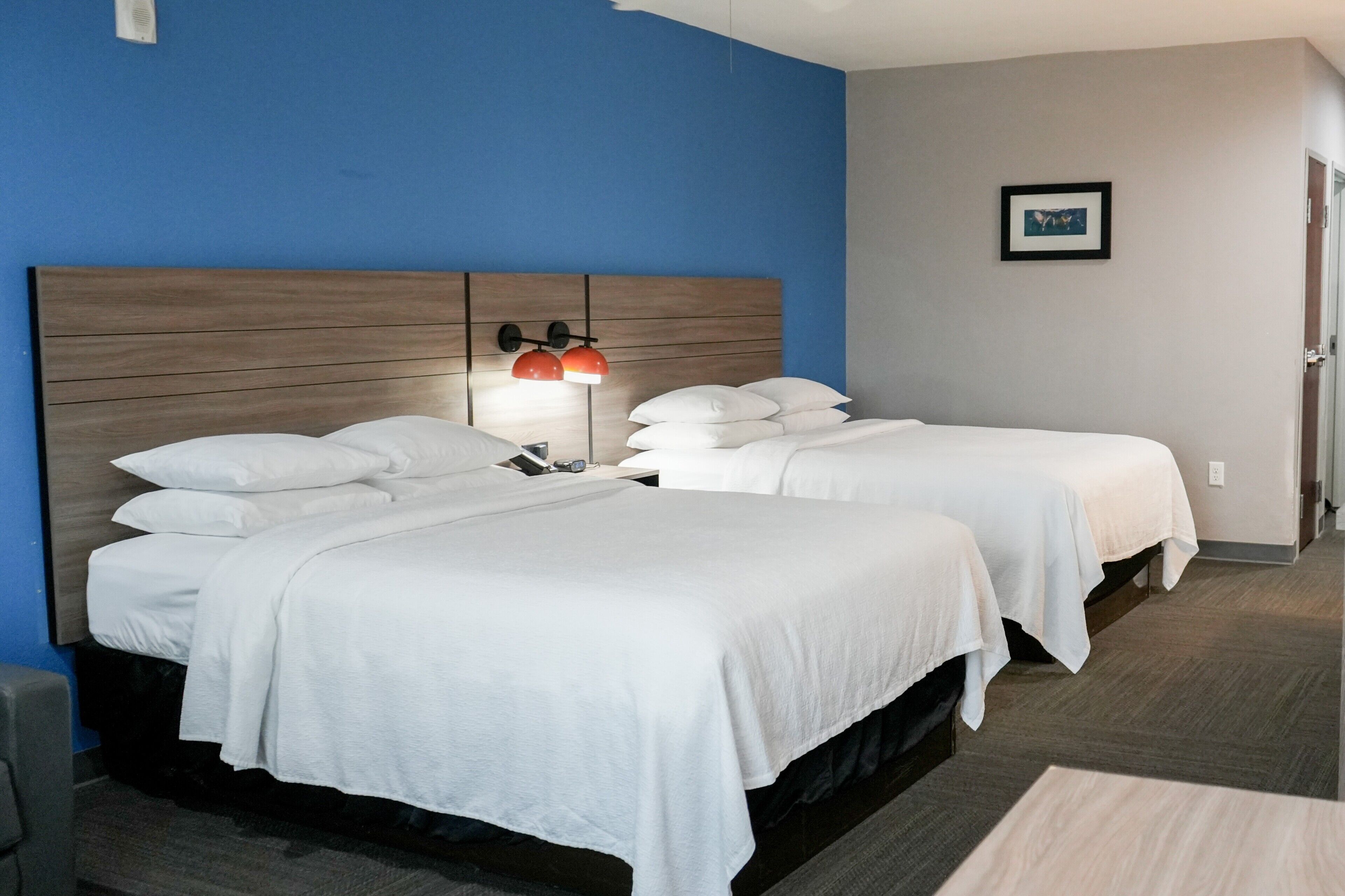 A hotel room with two beds, a wooden headboard, a nightstand, a red lamp, and a framed picture on the wall.