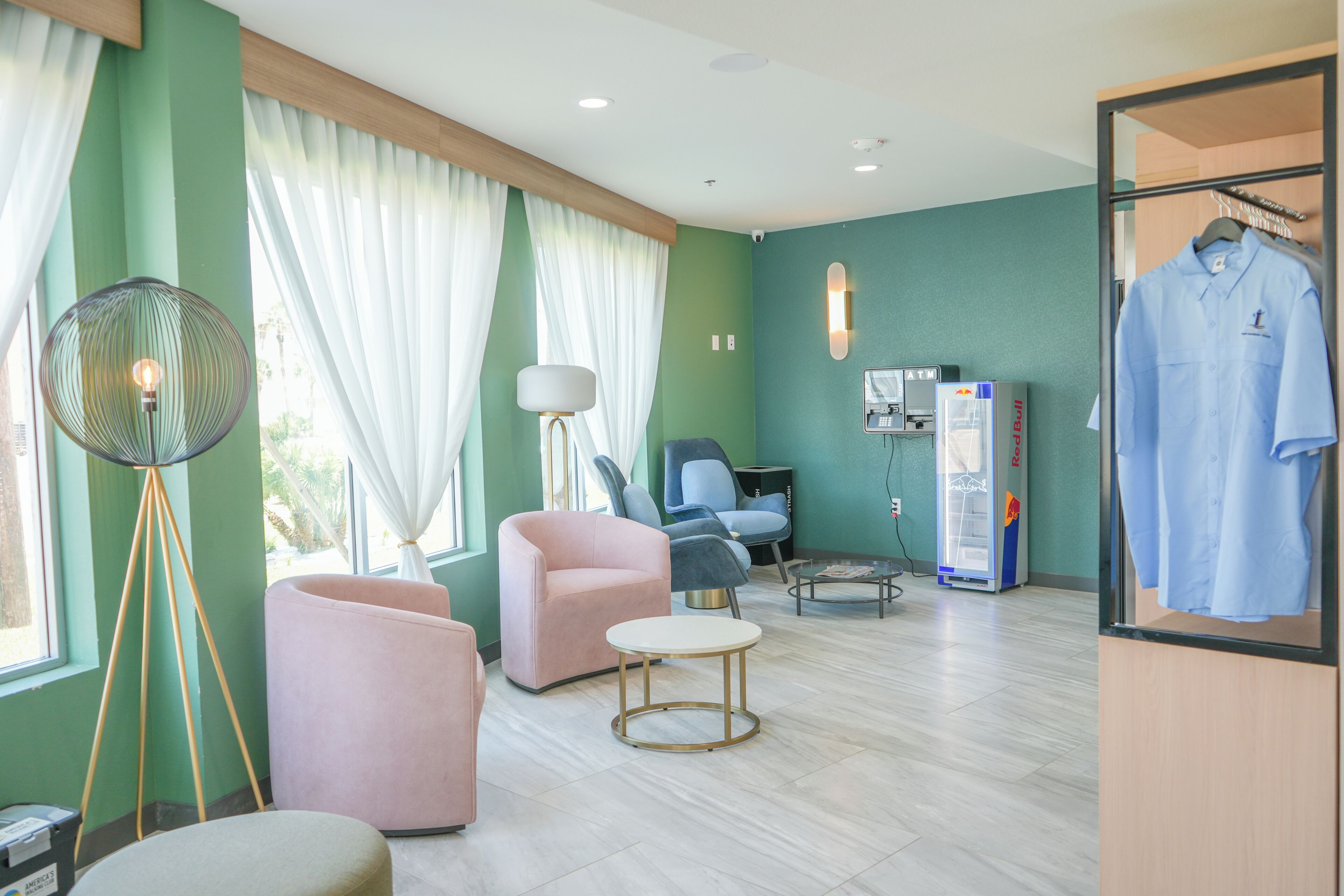 Une chambre d’hôtel avec des murs verts, une grande fenêtre avec des rideaux blancs, un fauteuil rose, un fauteuil bleu, une petite table ronde, un lampadaire, une télévision et un portant à vêtements.
