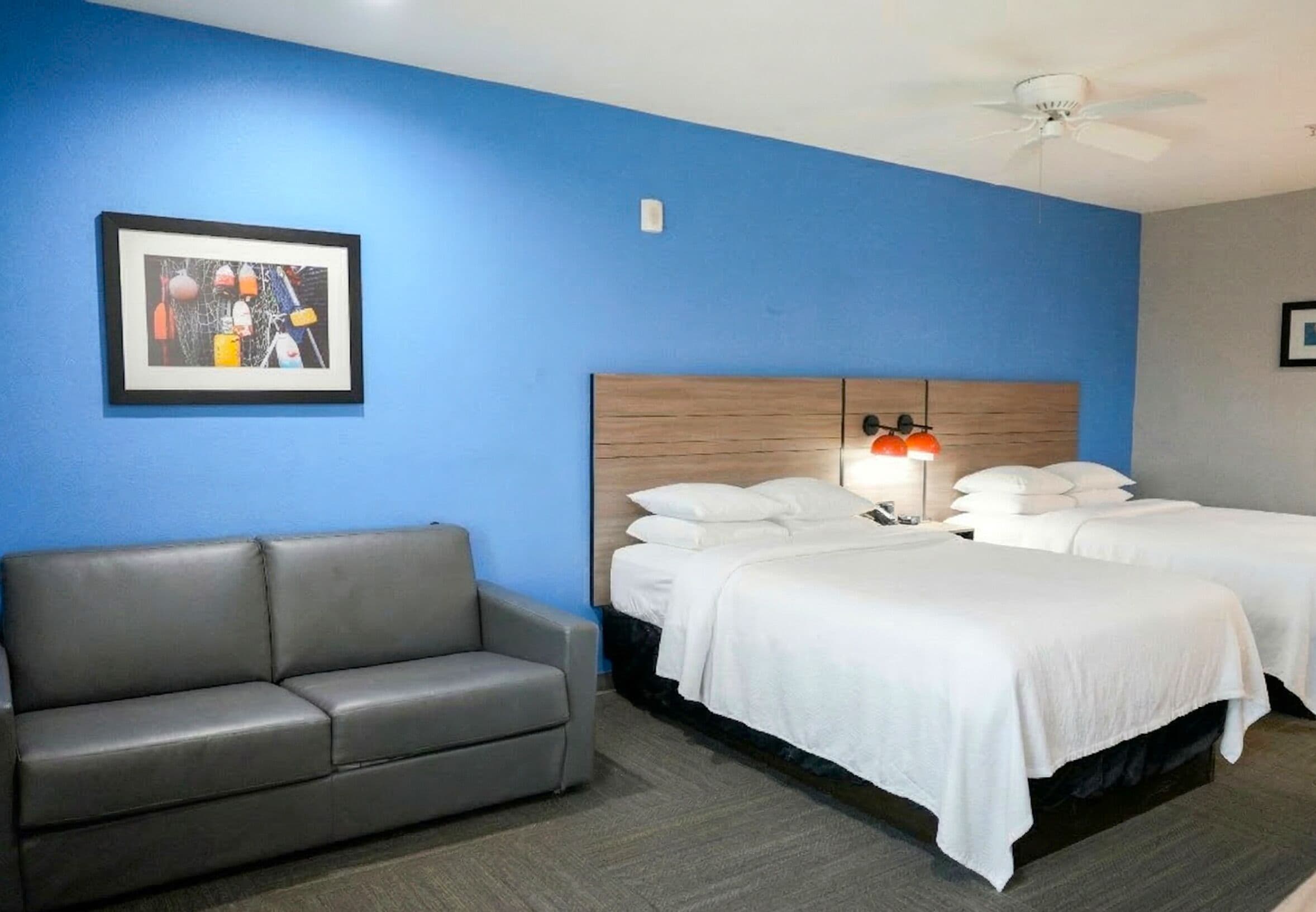 A hotel room with a blue wall, a gray sofa, two beds with white bedding, a wooden headboard, and a ceiling fan.