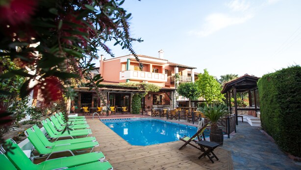 A pool area with lounge chairs, a wooden deck, and a building with outdoor seating. - Ammon Garden Hotel (Kassandra)