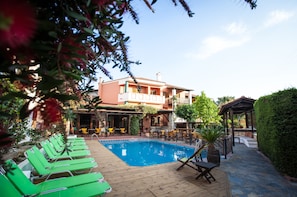 A pool area with lounge chairs, a wooden deck, and a building with outdoor seating. - Ammon Garden Hotel (Kassandra)