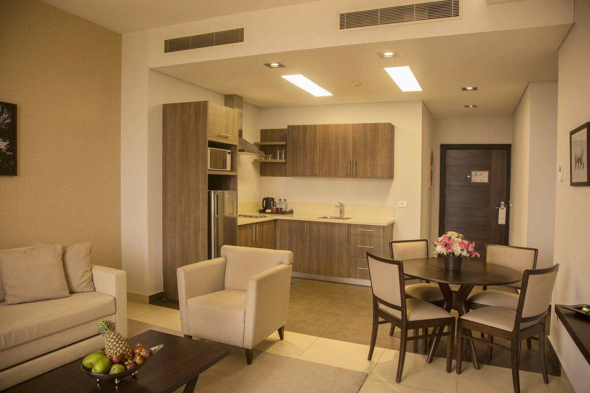 Un salon moderne comprenant un canapé, un fauteuil, une table basse et une table à manger avec des chaises. On aperçoit une cuisine à l’arrière-plan, avec des meubles et des appareils électroménagers en bois.