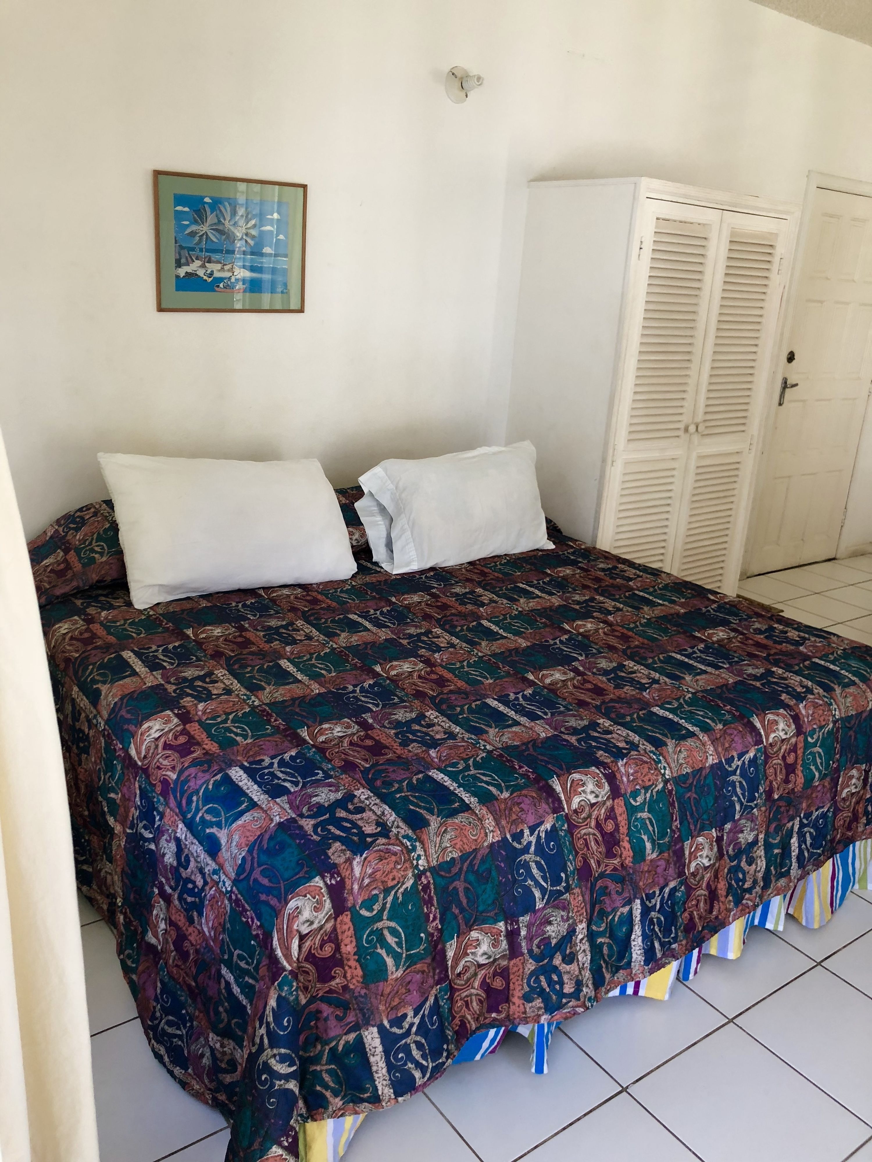 A bedroom with a bed, two pillows, a colorful quilt, a wardrobe, and a painting on the wall.