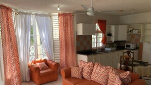 A living room with orange furniture, a sofa, two armchairs, a coffee table, and a kitchen area with white cabinets and a microwave.