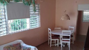 A room with a white sofa, a dining table with chairs, a hanging light fixture, and a window with blinds.