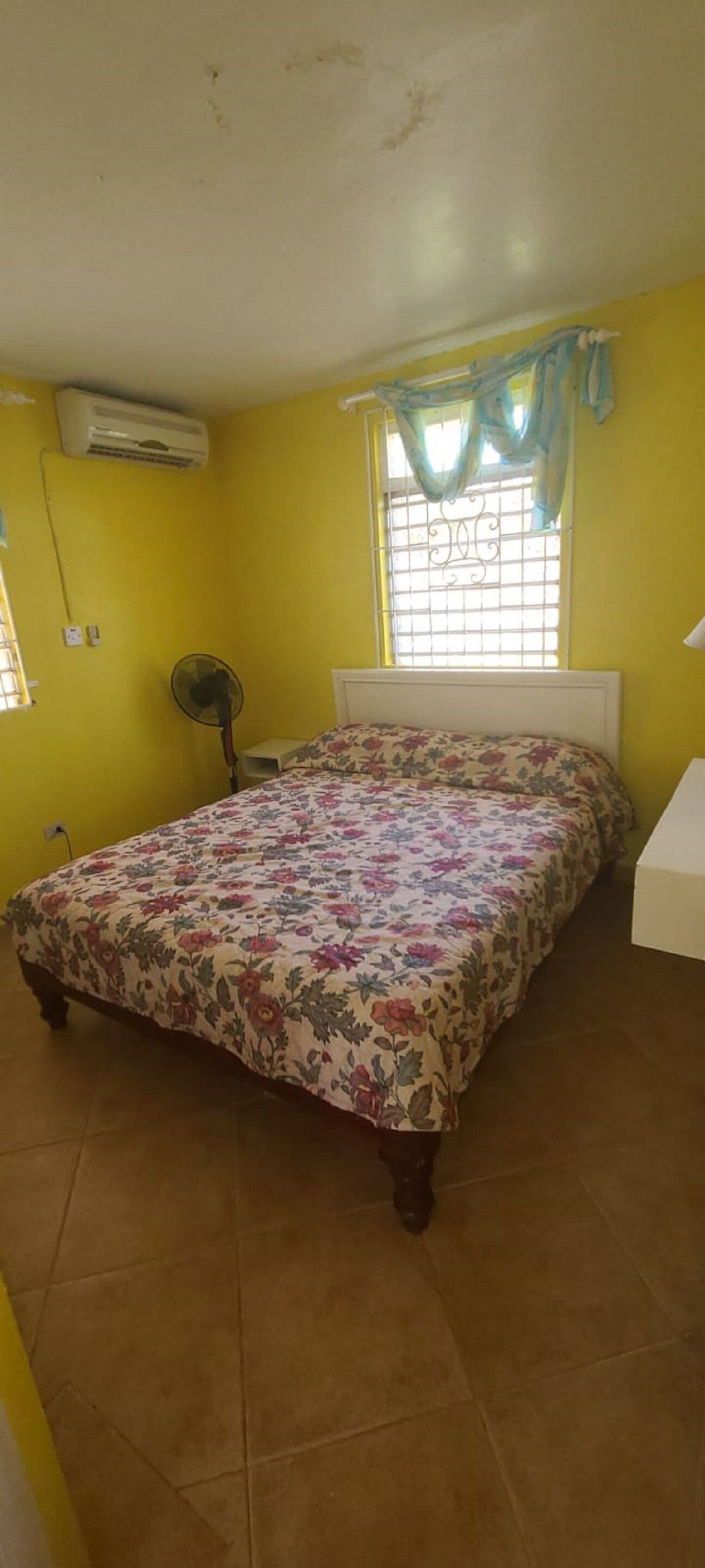 A bedroom with a floral bedspread, an air conditioning unit, a window with a curtain, and a bedside table.