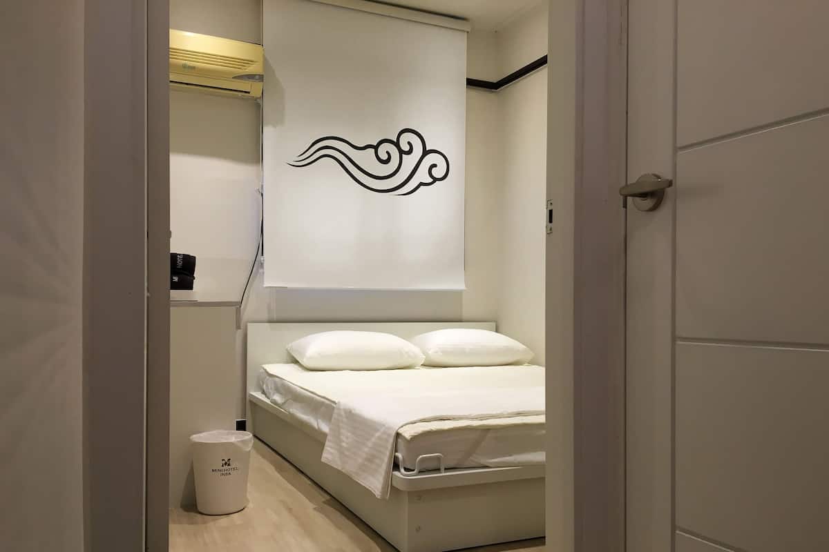Une chambre d’hôtel comprenant un lit, un climatiseur fixé au mur, une poubelle et un mur orné d’un motif décoratif.