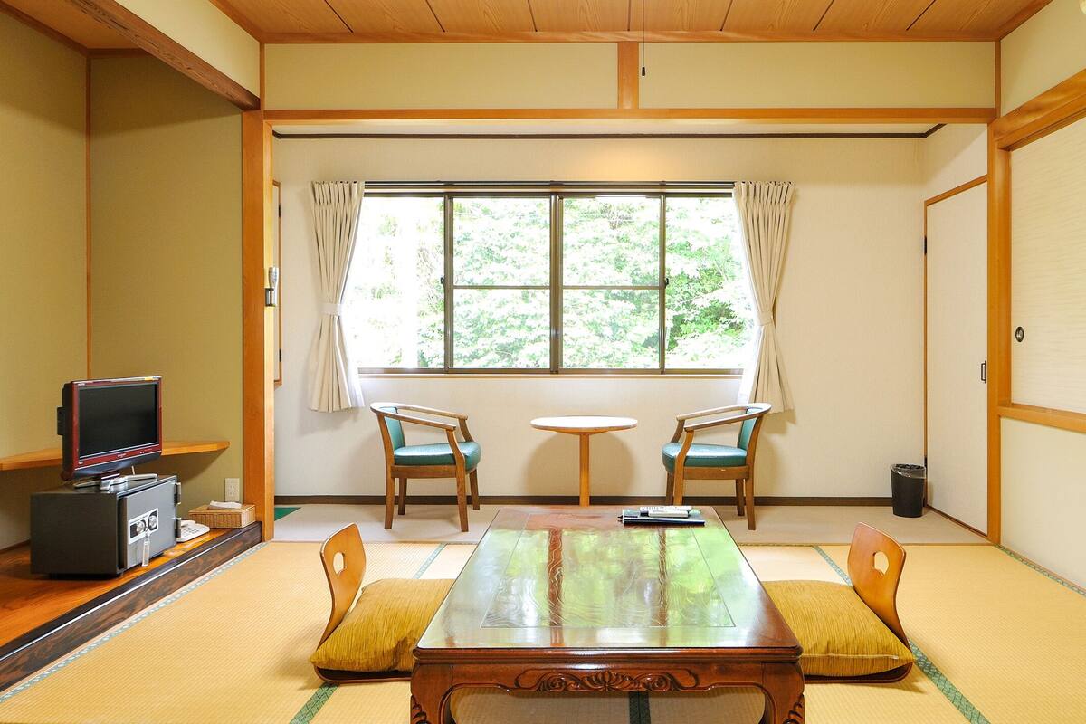 Une pièce de style japonais traditionnel, avec un sol recouvert de tatamis, une table basse, deux chaises, un téléviseur et une fenêtre avec des rideaux.