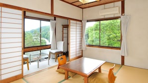 Una habitación japonesa tradicional con suelo de tatami, una mesa de madera, una silla y vistas a la vegetación exterior.