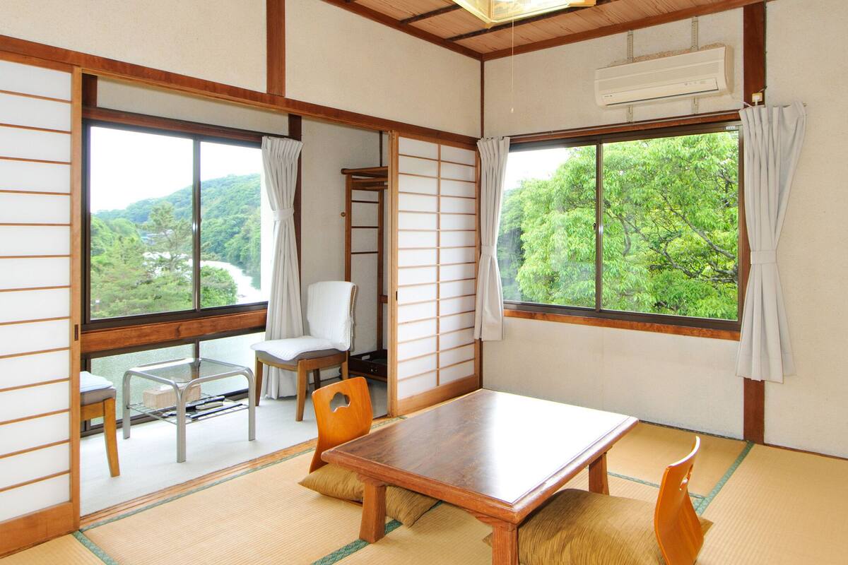 Une pièce de style japonais traditionnel, avec un sol recouvert de tatamis, une table en bois, une chaise et une vue sur la verdure à l’extérieur.