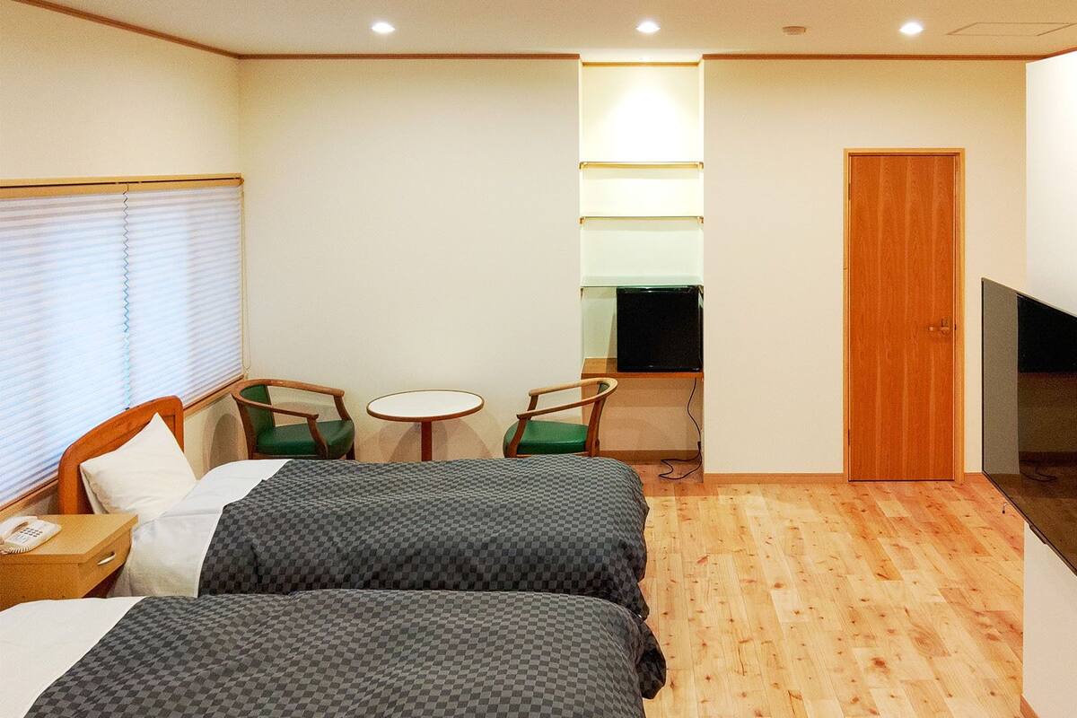 Une chambre d’hôtel avec deux lits, une télévision, un bureau avec une chaise, une petite table et une fenêtre avec des stores.