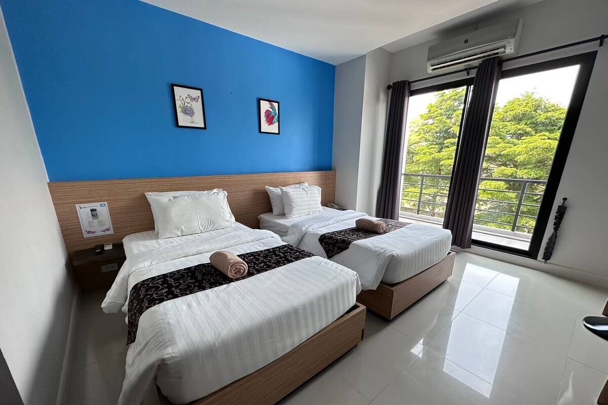 A hotel room with two beds, a blue accent wall, and a large window with a view of greenery.