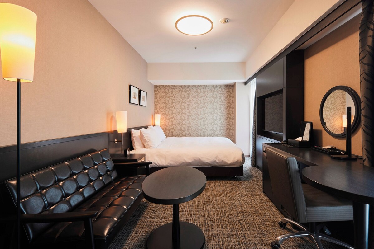 A hotel room with a bed, a sofa, a desk, a chair, and a TV.
