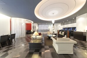 A modern hotel lobby with a checkered floor, a central seating area with a coffee table, a sofa, and a dining table with chairs. There are bookshelves and a reception desk with a counter and chairs. - Richmond Hotel Nagasaki Shianbashi (Nagasaki)