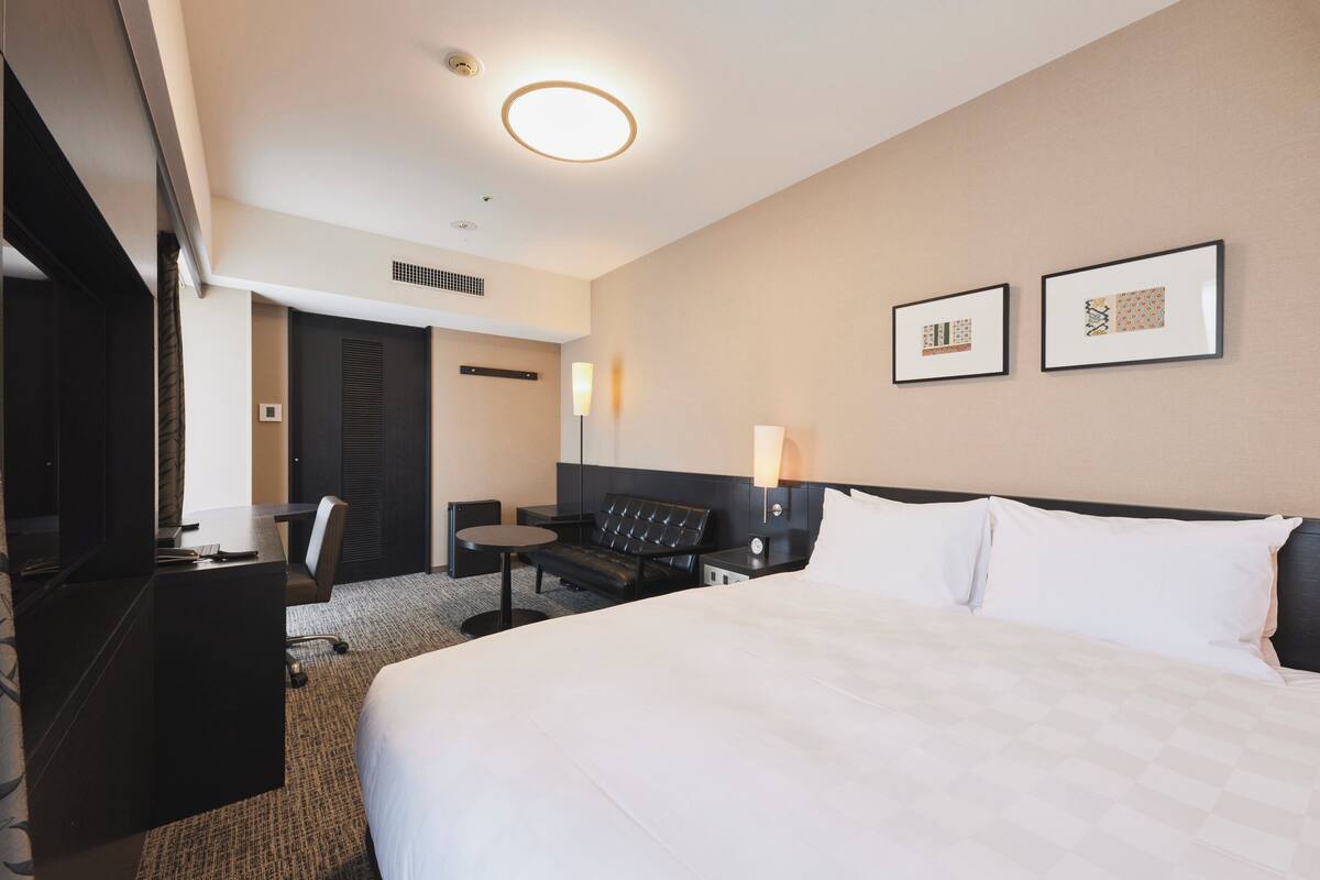 A hotel room with a large bed, a desk, a chair, a small table, and two wall-mounted lamps.