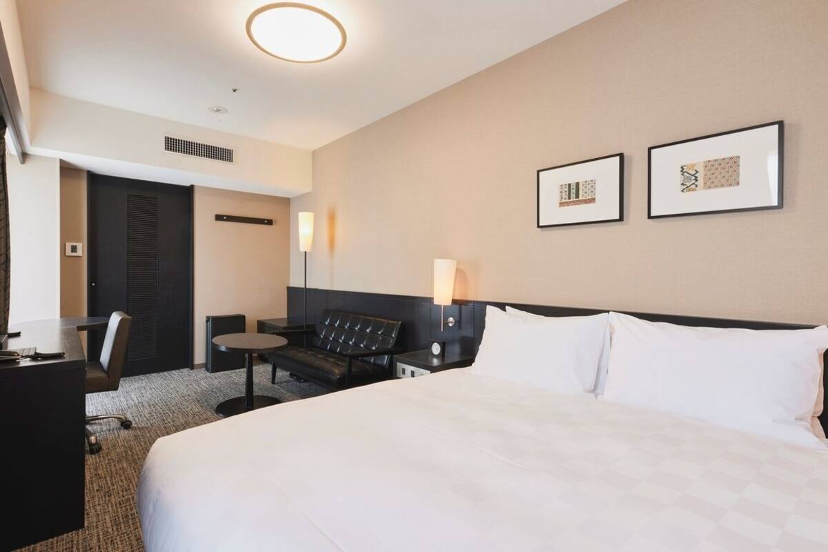 A hotel room with a bed, a desk, a chair, a small table, and two framed pictures on the wall.