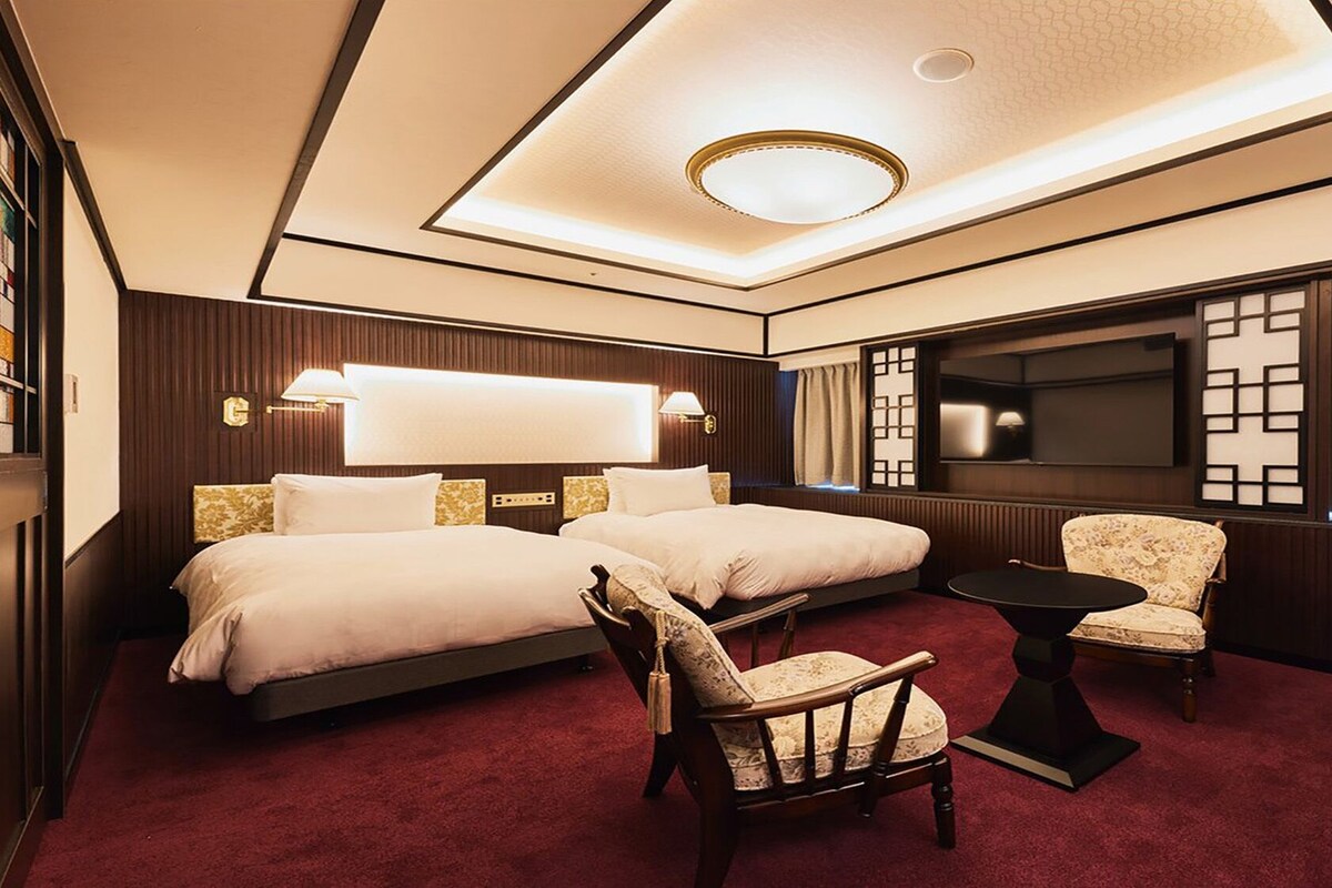 A hotel room with a large bed, a smaller bed, a seating area with a chair and table, a television, and a decorative wall panel.