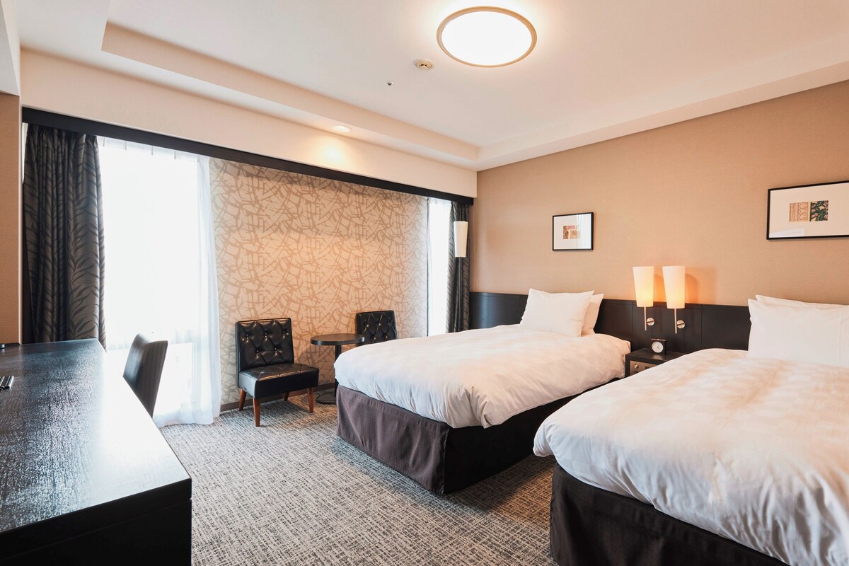 A hotel room with two beds, a desk, a chair, a small table, and a window with curtains.