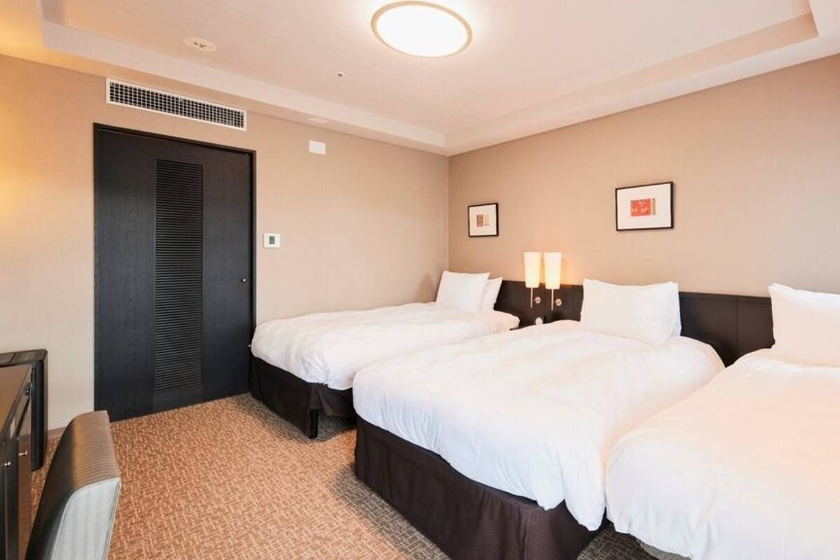 A hotel room with two beds, a dark-colored wardrobe, a small desk, and a wall-mounted light fixture.