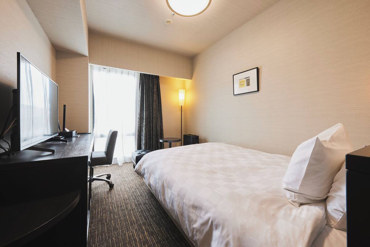 A hotel room with a bed, desk, chair, lamp, and a framed picture on the wall.