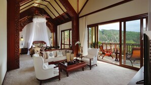 A spacious living area with a large sofa, armchairs, and a coffee table. There is a balcony with outdoor furniture and a scenic view of trees and hills.