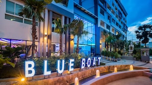 A hotel entrance with 'BLUE BOAT' sign, outdoor pool, palm trees, and a modern building facade. - Blue Boat Hotel JC (Pattaya)
