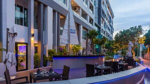An outdoor dining area with tables and chairs, a bar with a blue sign, and a sailboat model. - Blue Boat Hotel JC (Pattaya)
