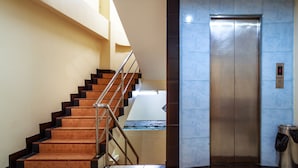 Um elevador moderno com acabamento metálico, uma escada com degraus de madeira e corrimãos de metal, e uma parede com parte revestida em azulejos azuis.