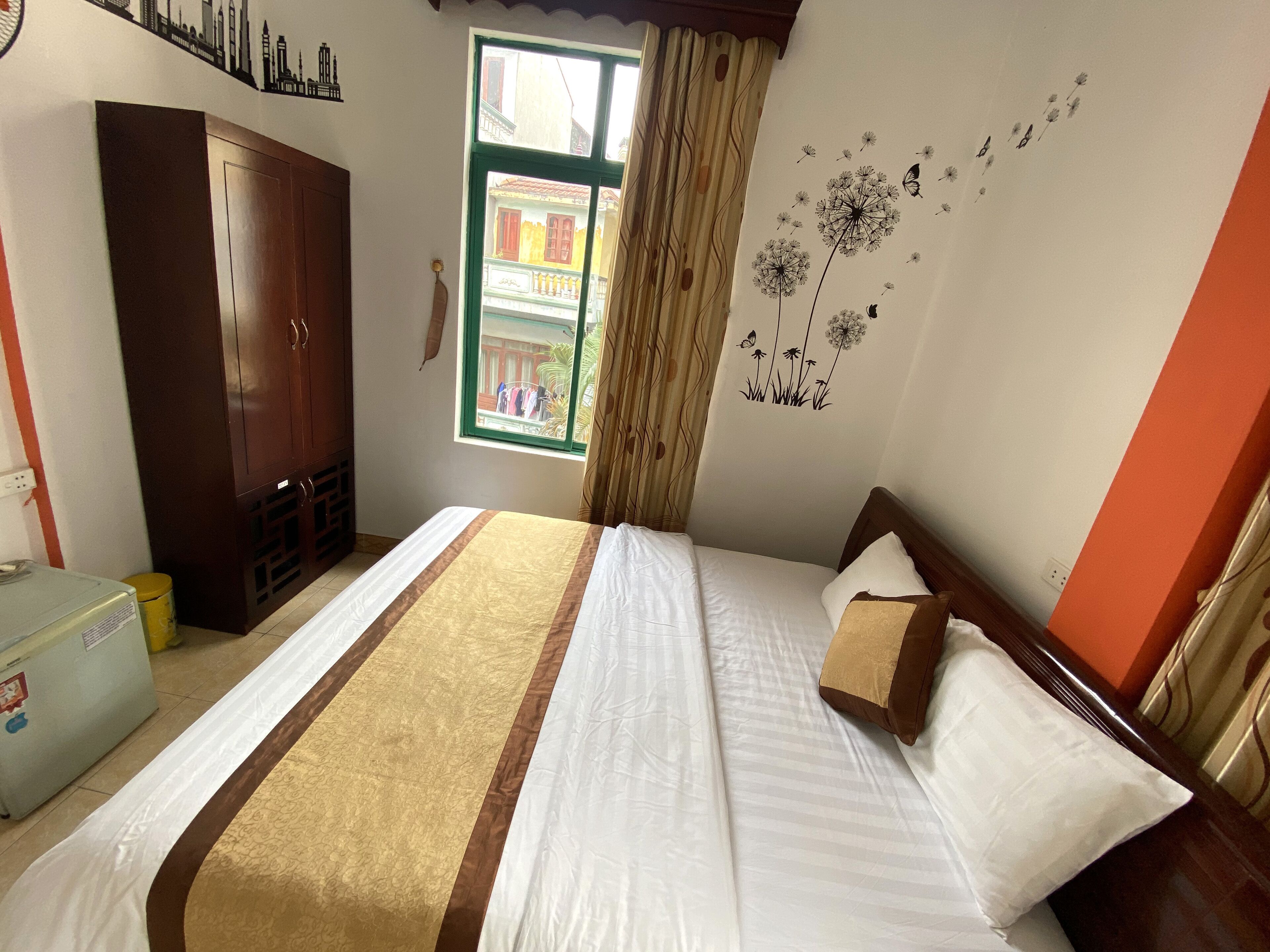 A neatly made bed with a striped bedspread, a wooden wardrobe, a window with a view of buildings, and a wall with a dandelion decoration.
