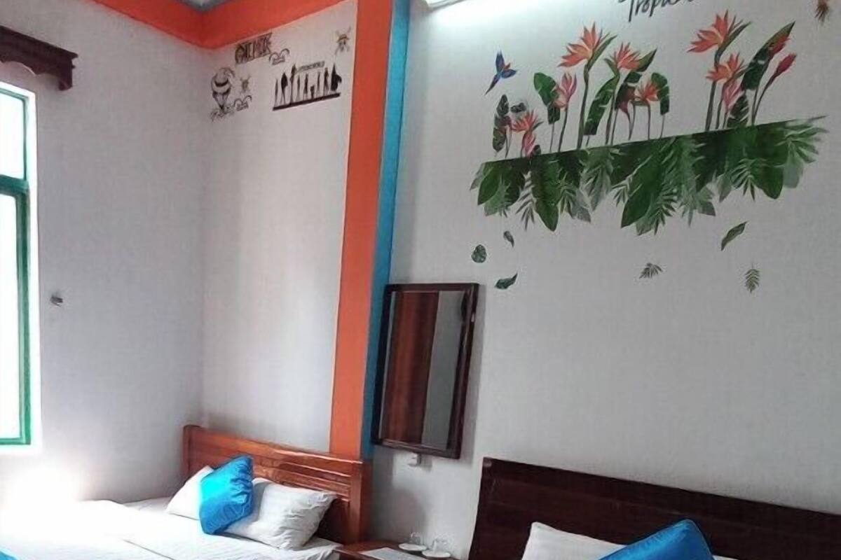 Une chambre d’hôtel avec deux lits, chacun agrémenté de draps blancs et bleus, d’une tête de lit en bois et d’une fresque murale représentant des fleurs et des oiseaux tropicaux.