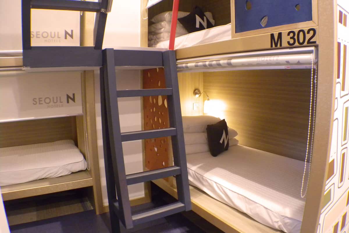Une chambre avec un lit superposé, une échelle, un oreiller orné de la lettre « N » et une enseigne indiquant « SEOUL N HOTELS ».