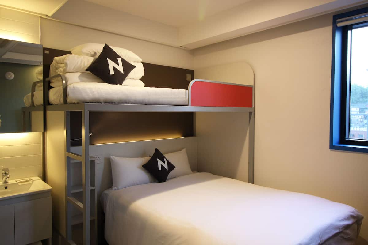 Un lit superposé avec une tête de lit rouge, un lavabo blanc et une fenêtre donnant sur des immeubles.