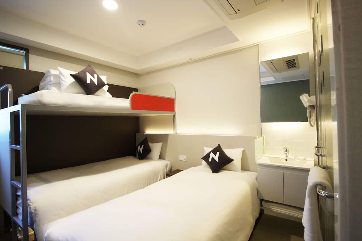 Une chambre d’hôtel avec deux lits simples, un lit superposé, une salle de bain avec un lavabo et un miroir.