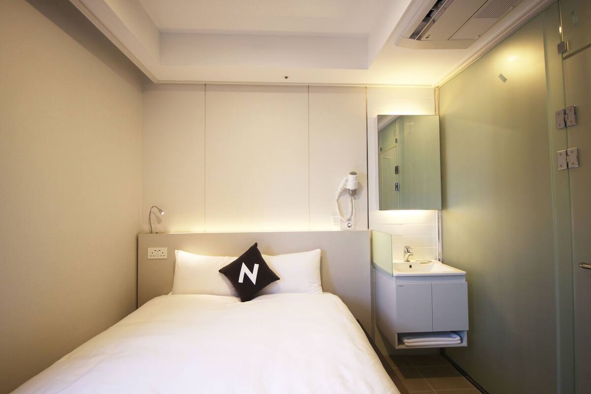 Une chambre d’hôtel comprenant un lit, une table de chevet avec une lampe, un miroir et une petite salle de bain avec un lavabo et un porte-serviettes.