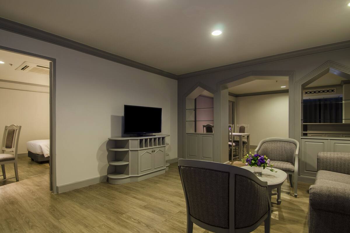 A modern living room with a flat-screen TV, grey cabinets, a dining table with chairs, and a small kitchen area.