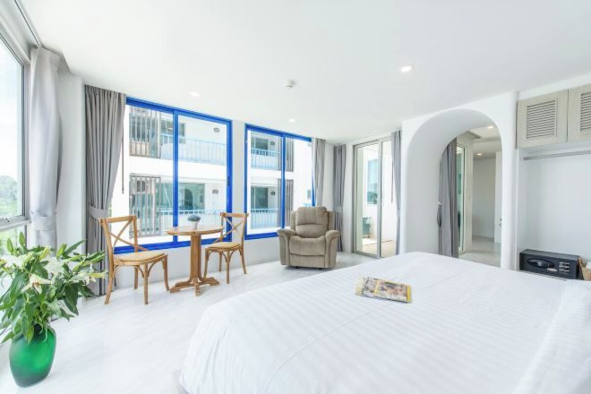 A modern hotel room with a large bed, a dining area with a table and chairs, a sofa, and a view of the building's exterior through large windows.