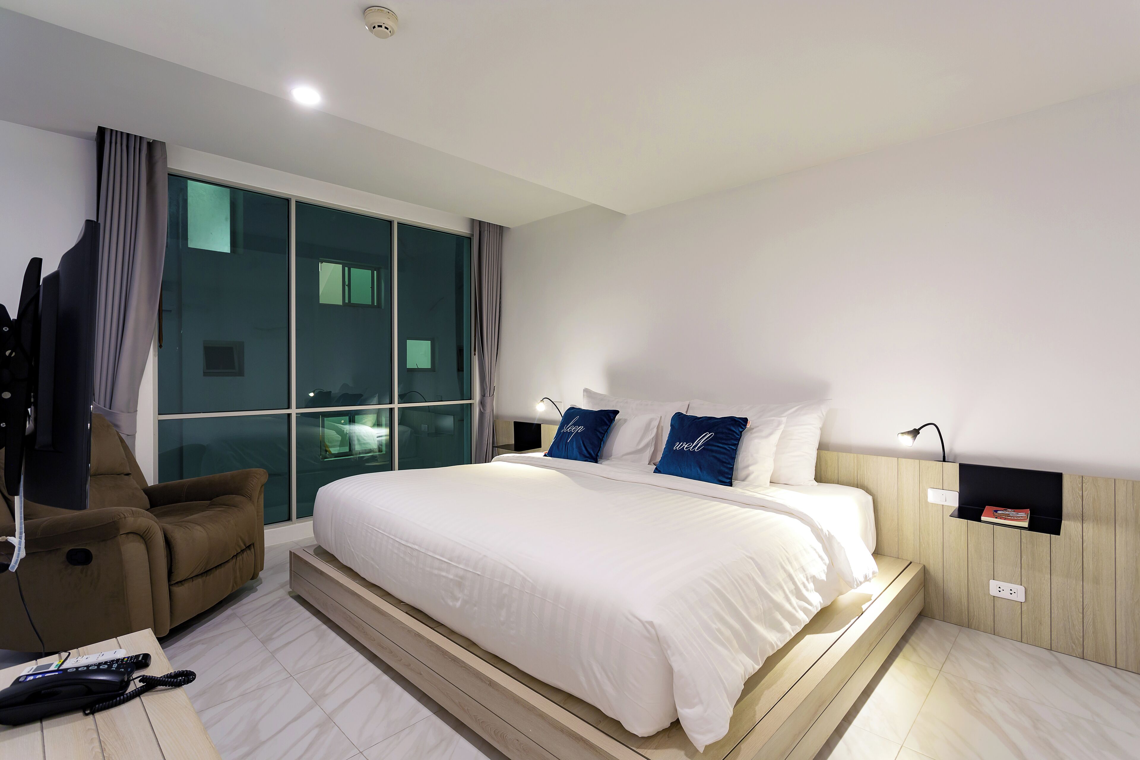A modern hotel room with a large bed, a reclining chair, a desk with a phone, and a view of the outside through a large window.