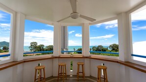 Ein Balkon mit drei Holzschemeln, einem Deckenventilator und großen Fenstern mit Blick auf ein hohes Gebäude und ein Gewässer.