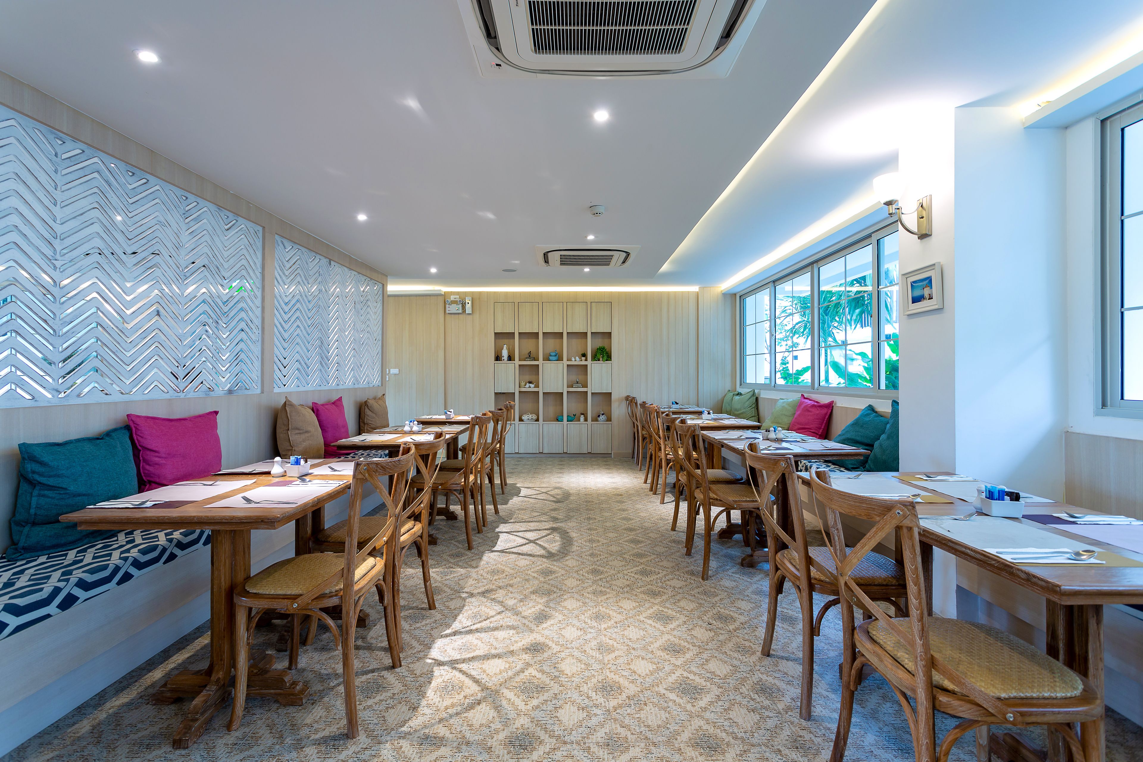 A spacious dining area with wooden tables and chairs, a patterned carpet, and a large window providing natural light.