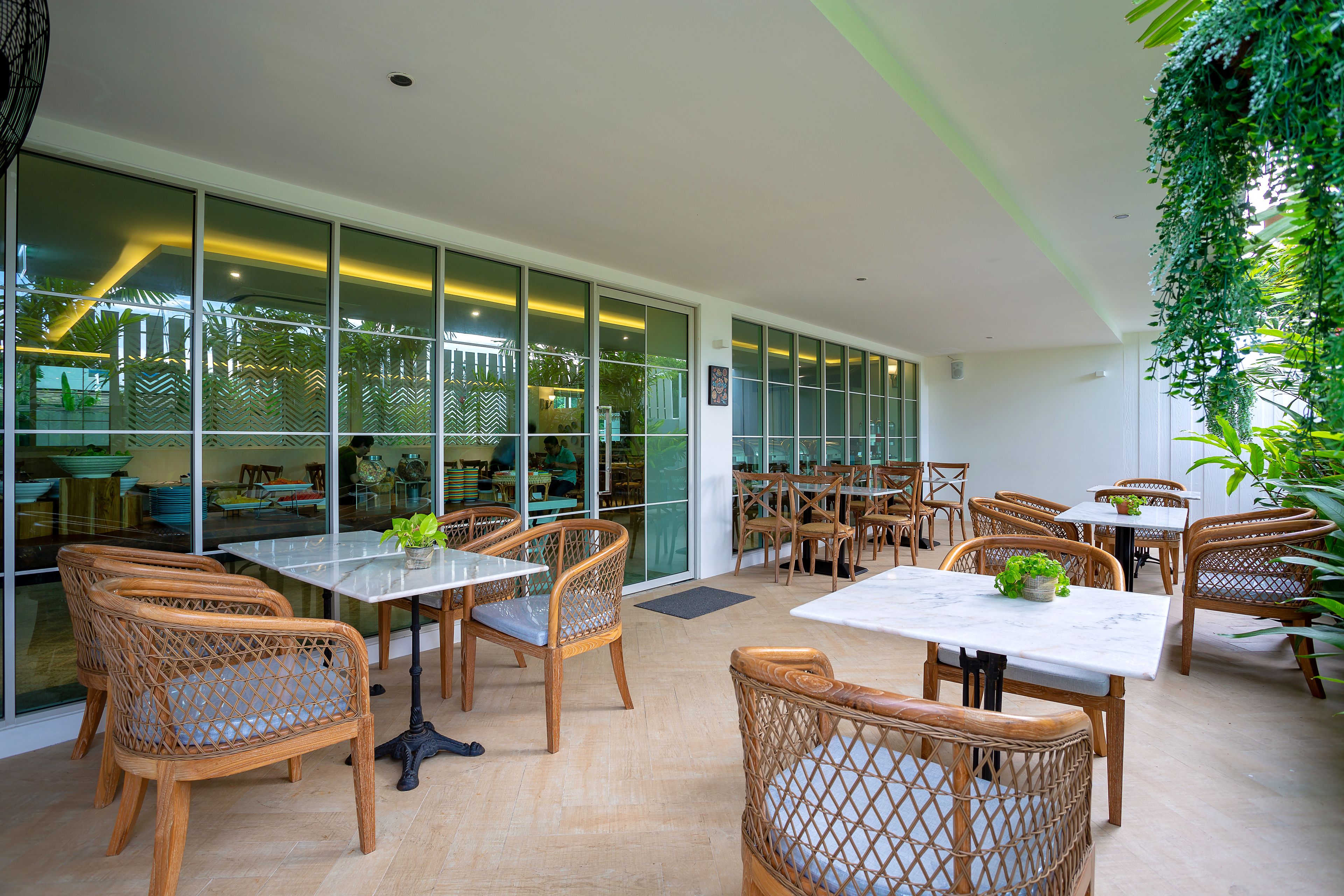 A modern indoor dining area with wicker chairs and tables, large windows, and indoor plants.
