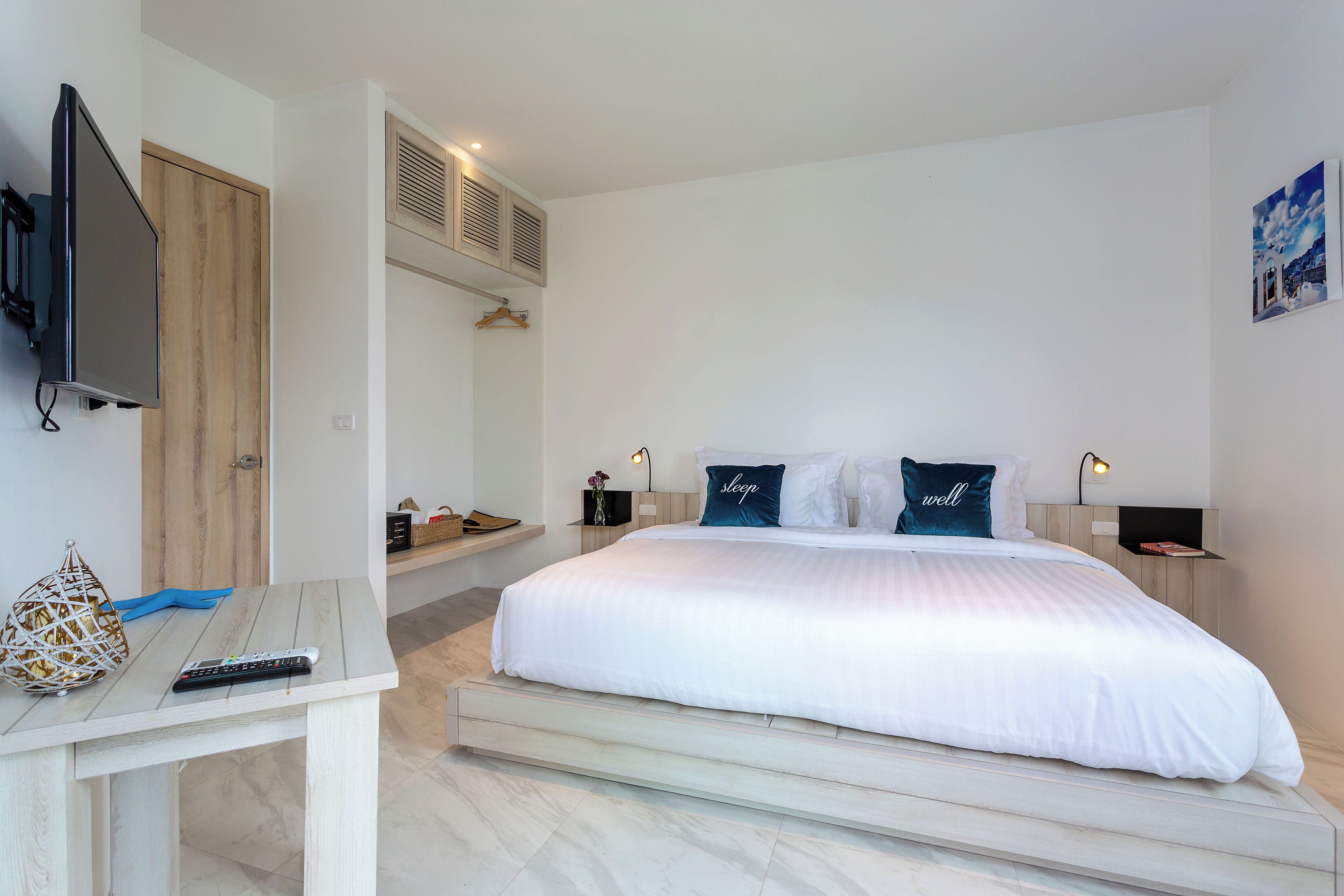 A modern bedroom with a large bed, a TV mounted on the wall, a desk with a remote control, and a small shelf with decorative items.