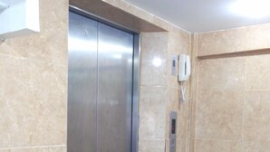 A modern elevator with a digital display, beige marble walls, and a plain white ceiling. - Al-Sana Hotel (Bangkok)