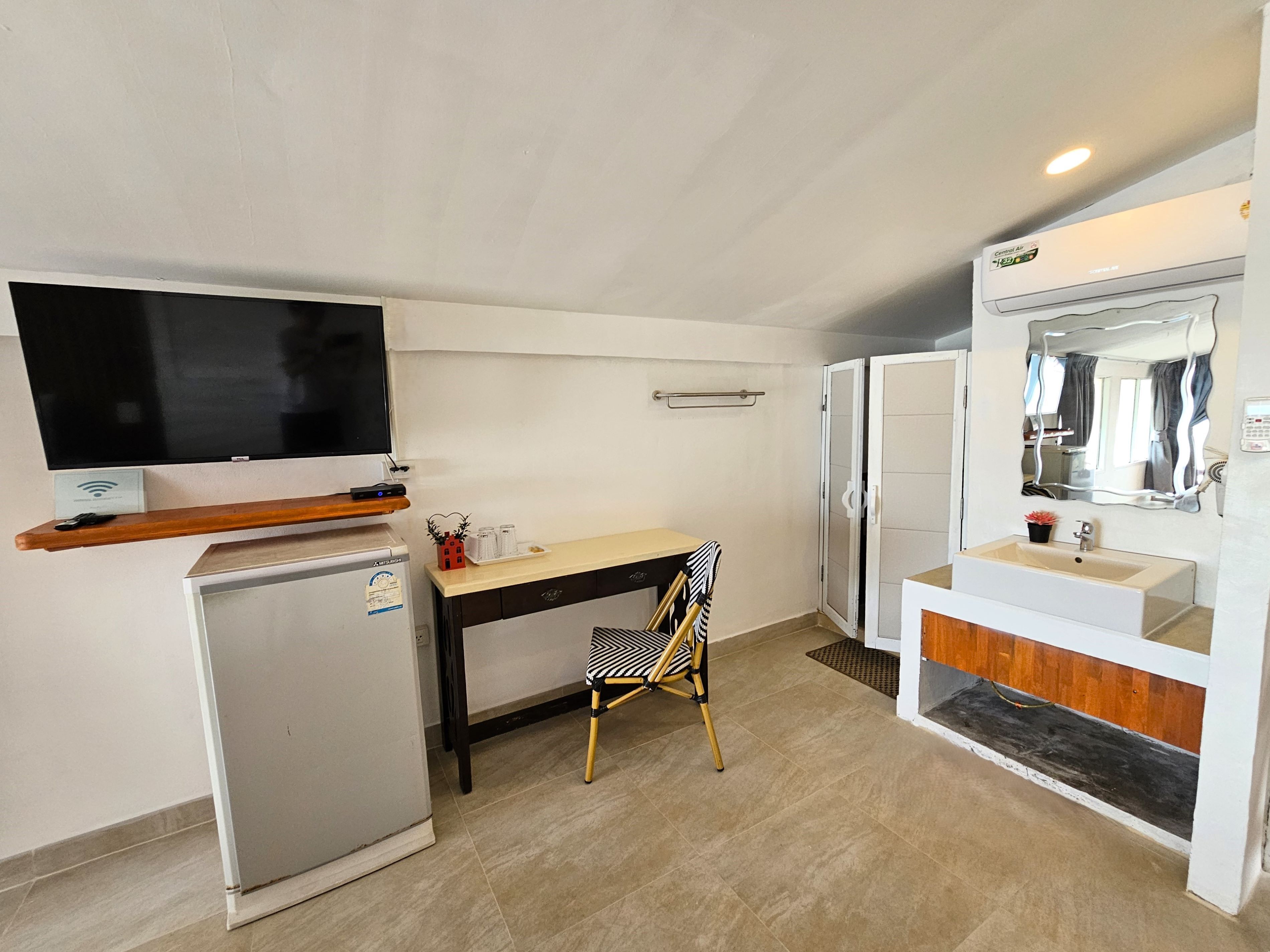 Een compacte hotelkamer met kitchenette, een kleine koelkast, een flatscreen televisie, een houten plank, een bureau met stoel, een badkamer met wastafel en spiegel, en een douche.