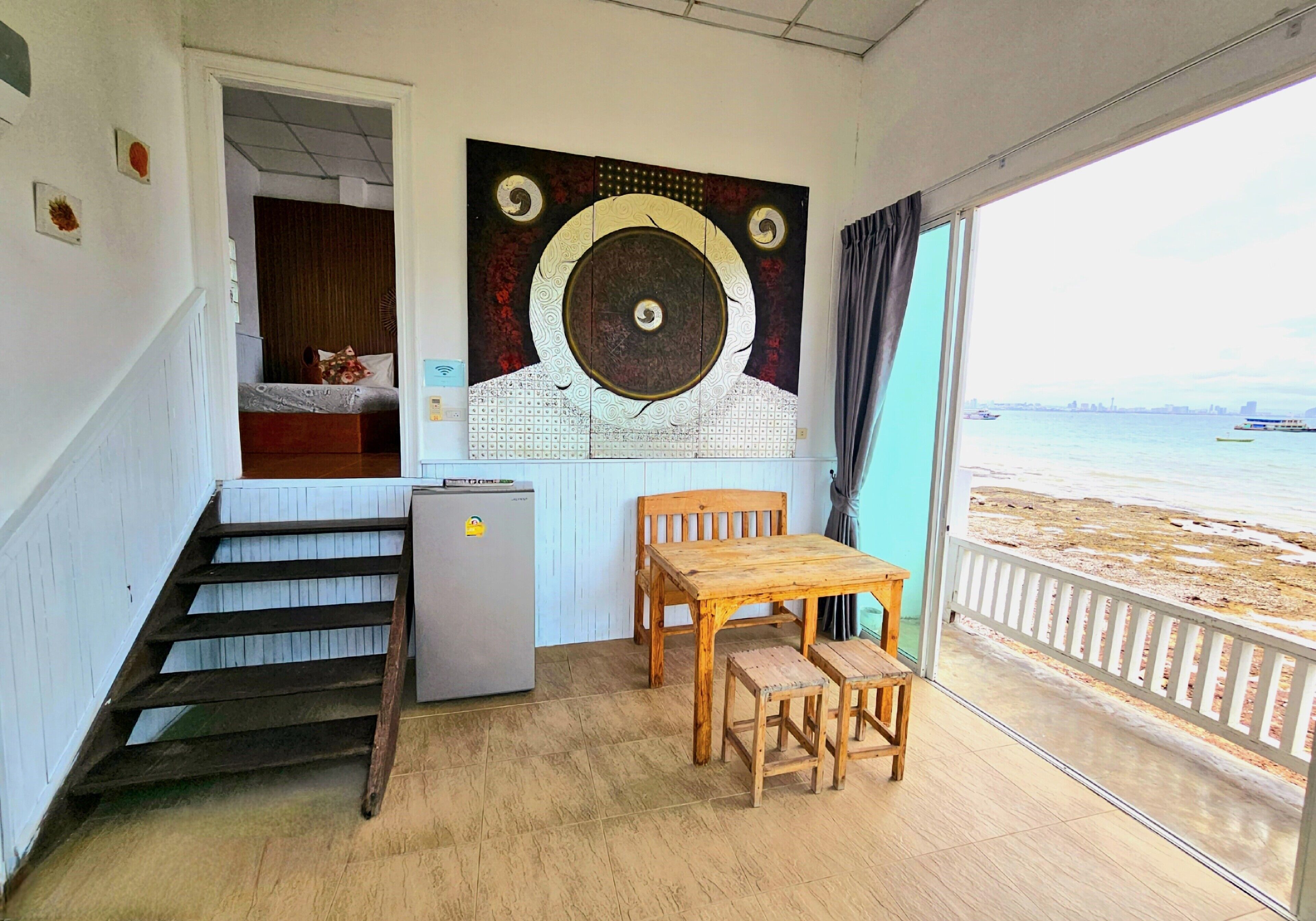 Une pièce avec une table et des chaises en bois, un petit réfrigérateur, un escalier menant à une chambre, et une grande fenêtre donnant sur la plage et les bateaux.